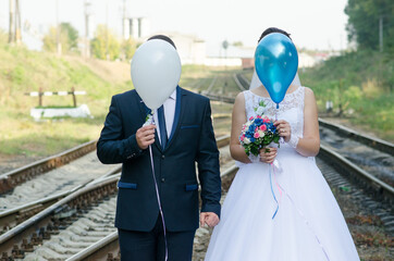 the bride with a bouquet and the groom with faces covered with balloons on the railway track, newlyweds with balloons on the railway track - Powered by Adobe