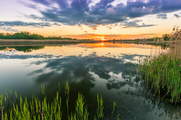 Fototapeta na wymiar Scenic view at beautiful spring sunset with reflection on a shiny lake with green reeds, bushes, grass, golden sun rays, water ,deep blue cloudy sky and glow on a background, spring evening landscape