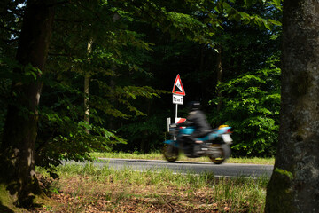 Motorradfahrer fährt kurvenreiche Waldstrecke, Warnschild zeigt Gefälle mit Prozentangabe.