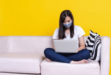 Fototapeta na wymiar Asian woman wearing medical mask and working from home use laptop computer while sitting on sofa over isolate yellow background