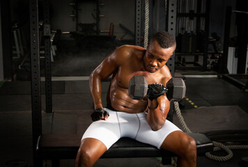 Selective focus of young African athlete sitting on a chair using dumbbell to build his biceps in a dark gym alone. African American man workouts by himself with dumbbell in shadow tone background
