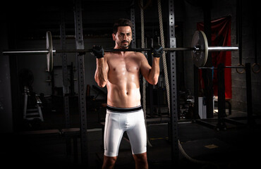 Fototapeta na wymiar Selective focus of young good looking Caucasian in a position to lift barbell in dark gym by himself. White muscle man workouts with barbell alone to build his body. Smoke background