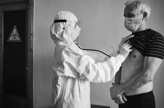 Doctor In Personal Protective Suit Examining Elderly Male Patient At Home Using Phonendoscope, Checking Chest, Standing At The Wall, Biohazard Sign On Door, Black And White, COVID-19 Concept