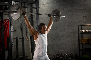 Naklejka na ściany i meble Selective focus of a young African American sport man workouts with barbells to build his body alone in a dark gym and little smoky background. Athlete plays with barbells to strengthen his muscles