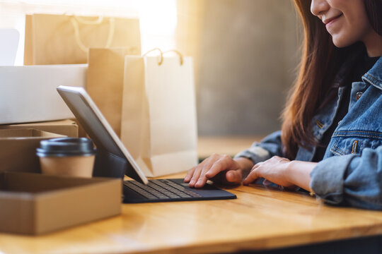 Closeup image of a young asian woman using tablet pc for online shopping with postal parcel box and shopping bags on the table at home