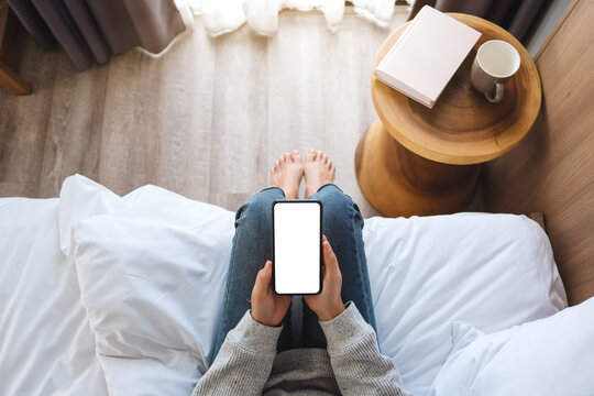 Top view mockup image of a woman holding mobile phone with blank desktop screen while sitting in bed room at home