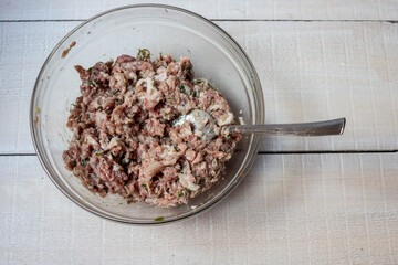 Cooking minced meat at home. Glass bowl and spoon with minced pork and beef with finely chopped greens on a white wooden background of boards close-up.