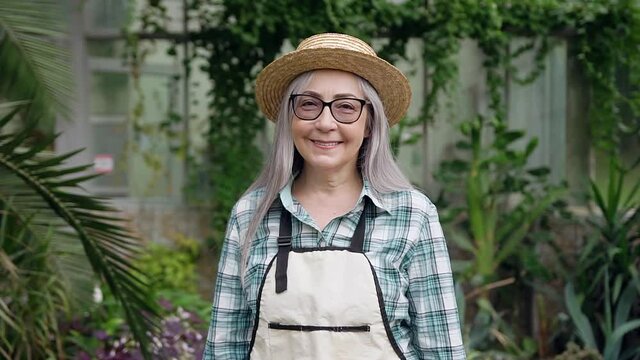 Beautiful smiling grey-haired woman in glasses wears hat and workwear with apron looking at camera in the wonderful orangery