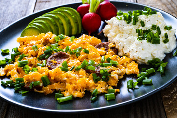 Delicious breakfast - scrambled eggs with fried sausages, cottage cheese, and avocado served on black plate on wooden table
