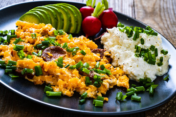 Delicious breakfast - scrambled eggs with fried sausages, cottage cheese, and avocado served on black plate on wooden table
