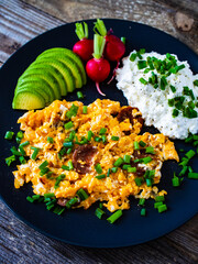 Delicious breakfast - scrambled eggs with fried sausages, cottage cheese, and avocado served on black plate on wooden table
