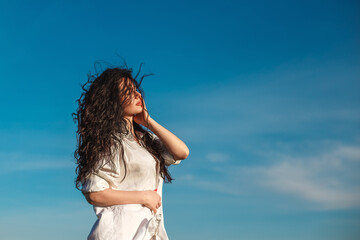 curly brunette girl in a white shirt on a background of blue sky. the wind blows long hair. merging...