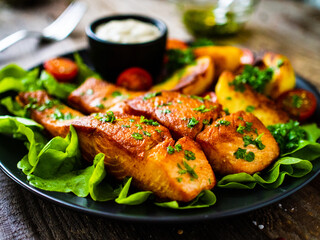 Fried salmon steak with potatoes and vegetables served on black plate on wooden table
