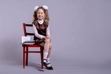 A schoolgirl in a school uniform and bows on her head sits on a chair, on a gray background, copy...