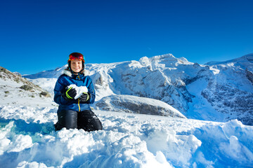 Fototapeta na wymiar Smiling teenage girl in blue outfit mask and helmet show love concept holding snow shape in heart standing on knees with mountain peak on background