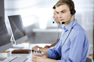 Two young men in headset, sitting at the desk in the modern office, listening to the clients. Call center operators at work