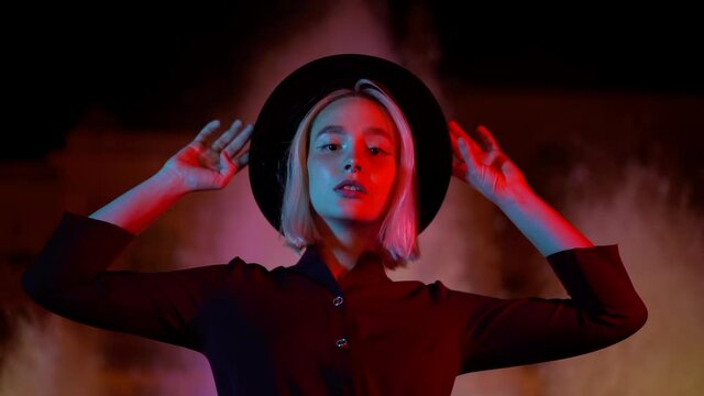 Portrait of millennial hipster woman with blond hairstyle standing on neon fountain background. City at night. Hat, nose piercing. Beautiful attractive girl. Slow motion.