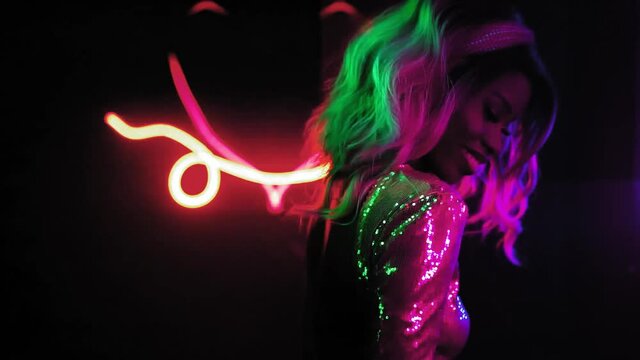 Sexy African-American blondie dances in disco shutter light herself in neon figures and in a silver sequin dress on a black background. Neon concept. Prores 422.