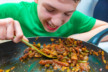 The boy eats fatty fried meat from a pan.