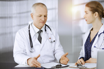 Elderly doctor and his female colleague discussing current disease therapy while sitting at working...