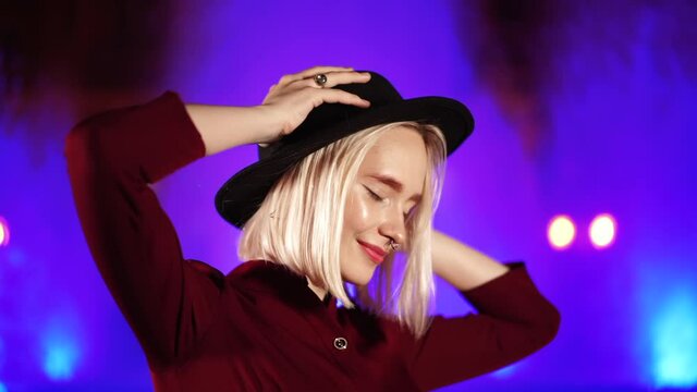 Portrait of charming charismatic hipster woman with blond hairstyle standing on neon fountain background and smiling. City at night. Hat, nose piercing. Beautiful attractive girl. Slow motion.
