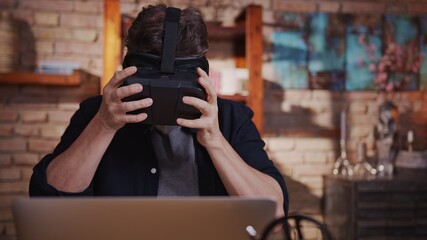 Casual man man playing video game on virtual reality googles at home sitting at desk with laptop computer.