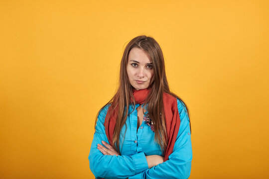 Standing and cross her arms on chest. Young attractive woman with brown hair, eyes, decoration on blue shirt, yellow background