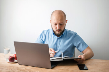 Bald young man with beard reading encyclopedia book and working online on his laptop at the wooden table in home office, white wall as background. Business, freelance or study concept