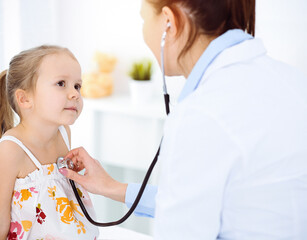 Doctor examining a child by stethoscope in sunny clinic. Happy smiling girl patient dressed in bright color dress is at usual medical inspection