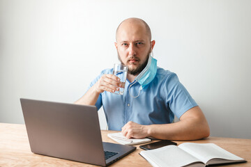 Bald business man with beard takes off surgical medical mask on face as protection against coronavirus, covid-19, and drink water from glass, working online on laptop at office
