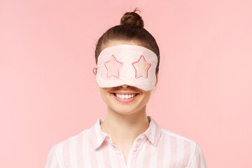 Young smiling teenage girl wearing pyjama and soft sleeping mask over eyes, isolated on pink background