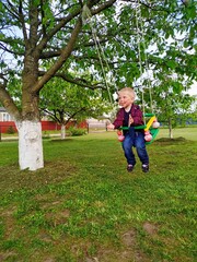 Fototapeta premium joyful kid dressed in a red plaid shirt and blue jeans swinging on a swing on the backyard. Happy moments