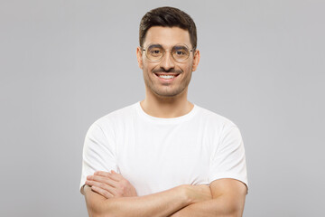 Young handsome smiling man wearing white t-shirt and eyeglasses, standing with arms crossed, feeling confident as professional, isolated on gray background