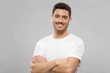 Portrait of young handsome guy in white t-shirt and wireless earphones holding arms crossed, feeling confident at work, isolated on gray background