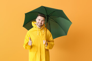 Young man in raincoat and with umbrella showing thumb-up on color background