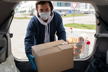 Middle-aged man in cloth mask and jacket standing at car and loading box into trunk after visiting store