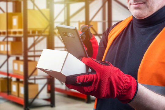 A man in work gloves on the background of the warehouse. The work of the storekeeper. A man with a smartphone and a cardboard box. Warehouse account. Warehouse activities.