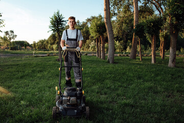 A young male gardener in overalls uses a lawn mower.