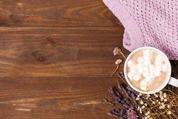 Hot chocolate or cocoa with marshmallows in a large white mug, a lilac knitted sweater and dry lavender flowers on a brown wooden background. The concept of cozy autumn. Top view, flat lay, copy space