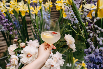 Female hand holding a glass with fresh homemade lemonade with lemon slice and strawberry, on a flower background in a garden.