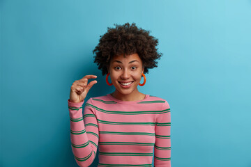 Some more, please. Cheerful smiling African American woman gestures small size with fingers, asks for little bit time, measures too small object, shows something minimum dressed casually stands indoor