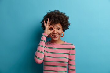 Portrait of funny cheerful Afro American woman makes okay gesture over eye, completely agrees with suggestion, smiles broadly, wears striped sweater, isolated on blue background, Everything is fine