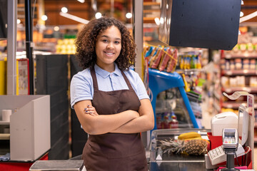 Girl in apron standing by cashbox in supermarket and crossing arms by chest