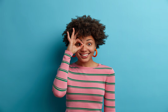 Portrait of funny cheerful Afro American woman makes okay gesture over eye, completely agrees with suggestion, smiles broadly, wears striped sweater, isolated on blue background, Everything is fine