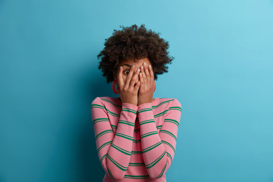 Afro American woman peeks through fingers, covers face with hands, dressed in casual striped sweater, afraids of something, hides face, isolated on blue background. People and fright concept