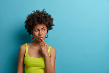 Serious looking dark skinned woman touches lips and looks thoughtfully aside, has troublesome thoughts, wears casual summer vest, needs think up plan, isolated on blue background, empty space