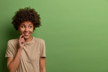 Obraz na płótnie Canvas Portrait of optimistic glad curly woman looks away with engaging toothy smile, expresses joy and happiness, wears casual beige t shirt, isolated over green background, copy space for promo or text