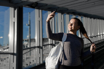 Young beautiful smiling girl makes selfie. Portrait of model in fashion sportswear on urban background on a sunny summer day.