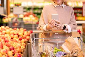 Mature female consumer in knitted beige sweater pushing cart with food products