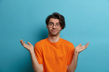 Fototapeta premium Clueless adult man says dont know, maybe, who cares, looks disinterested in question, has doubts, stands bewildered, spreads palms sideways, wears optical glasses and orange t shirt, isolated on blue